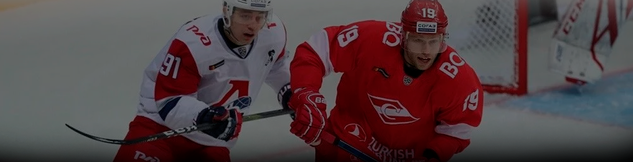 На изображении два хоккеиста в форме разных команд борются за шайбу на льду. Один из игроков в красно-белой форме, другой в полностью красной. Видна часть игровой площадки